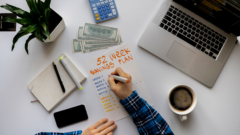 Male hand writing out a 52-week money savings challenge plan on desk with coffee, laptop, and plant