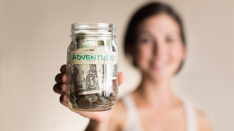 Woman holding jar with coins and dollar bills labeled "adventure" indicating light-lifting money saving challenges