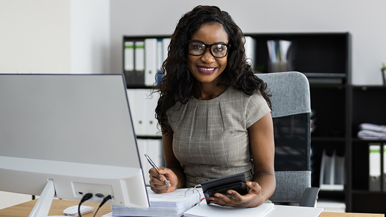 female accountant sitting at computer, holding calculator in front of a large binder