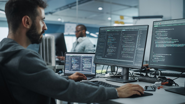 Male software engineer in front of laptop and two monitors, writing code