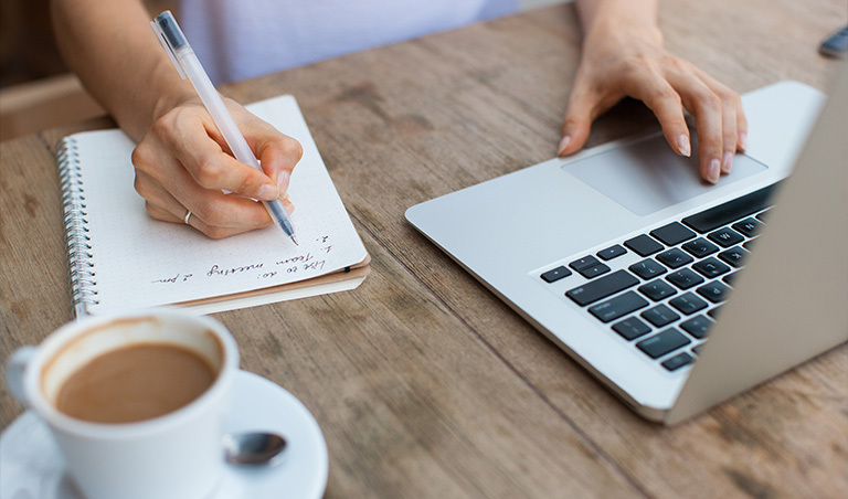 View of a woman's hands making a list of subscriptions on a notepad and working on a laptop