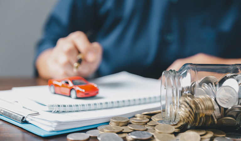 hands hovering over notepad with red toy car on top and jar of coins spilling out, implying buying a car